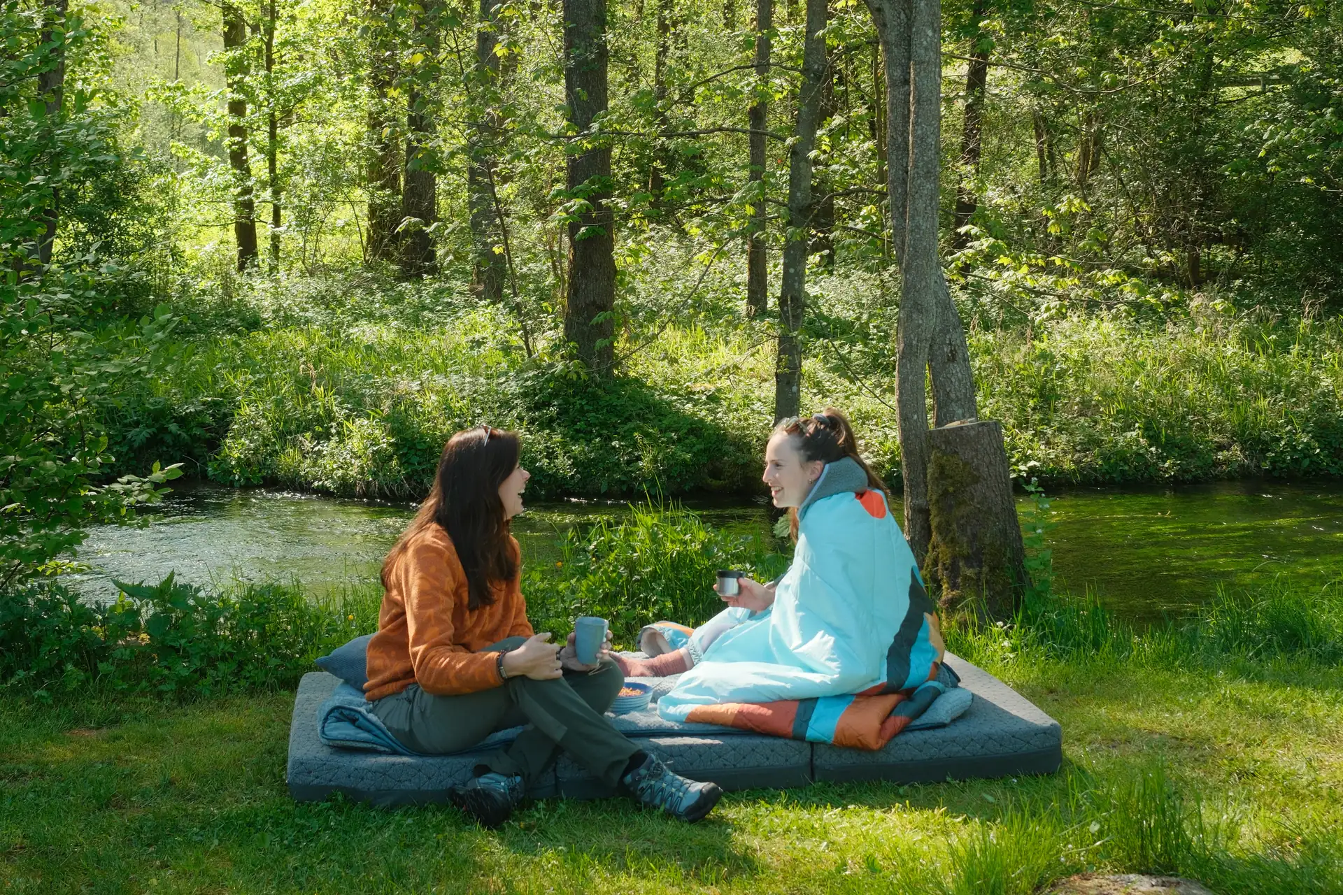 Im Einsatz, zwei junge Freundinnen sitzen auf einer Vanmade Original Matratze im gruenen an einem Bach